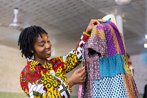 Smiling Ghanaian Dressmaker with locs hair measures a part of her colourful African pattern dress in her studio