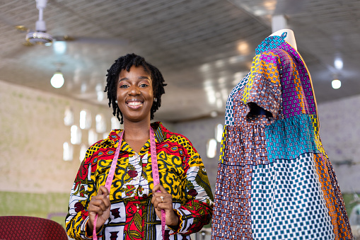 Successful African Fashion Dressmaker Entreprenuer standing by her Colorful African Print Pattern Dress with measuring tape around her neck.
