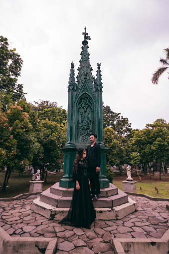 a couple of Asian teens standing near a spooky cemetery full of green trees in the afternoon