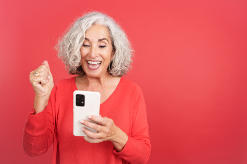 Studio portrait with red background of a mature woman celebrating while using the mobile phone