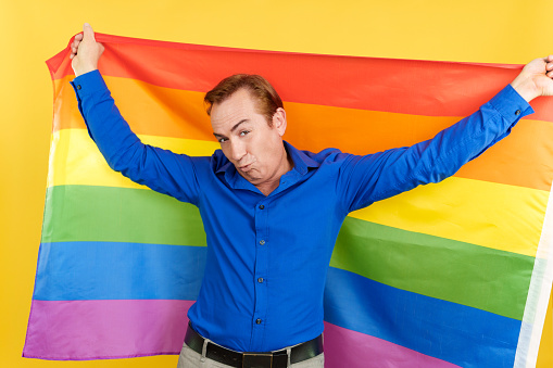 Happy mature man blowing a kiss raising a lgbt rainbow flag in studio with yellow background