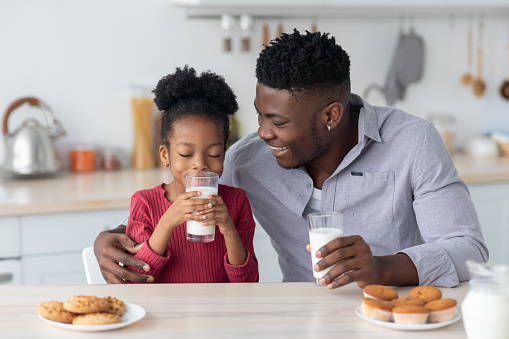 Healthy kids lifestyle, children nutrition concept. Cute little black girl drinking fresh milk, sitting at table with her cheerful father, eating cookies and cupcakes, child spending time with her dad