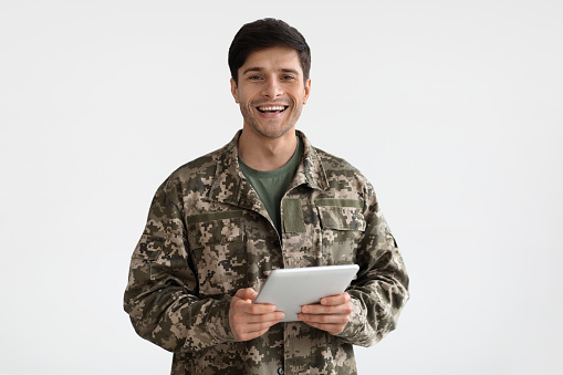 Smiling millennial man in camouflage uniform holding modern digital tablet, chatting with family, posing on white studio background, copy space. Modern technologies and military personnel concept