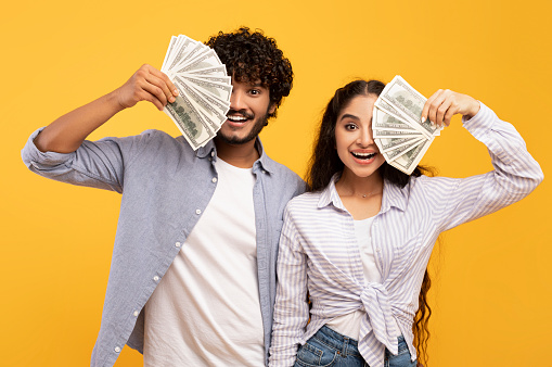 Rich millennial indian lovers hiding faces behind cash, holding bunches of dollars by faces and smiling over yellow studio background, enjoying prize, trading or gambling together
