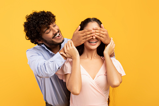 Romantic surprise concept. Portrait of happy indian guy covering his pretty girlfriend eyes from back, standing behind her, couple posing on yellow studio background, banner