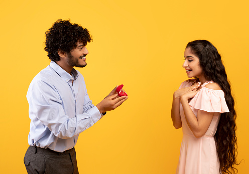 Marry me concept. Happy indian man proposing showing engagement ring box and asking surprised girlfriend to be his wife, standing over yellow background