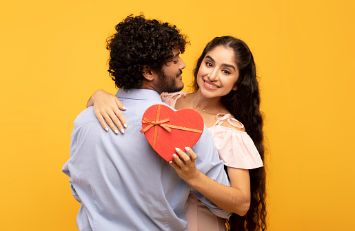 Young indian lovers hugging on yellow background, lady holding heart shaped gift box and smiling at camera. Loving couple celebrating Valentine's Day or anniversary together