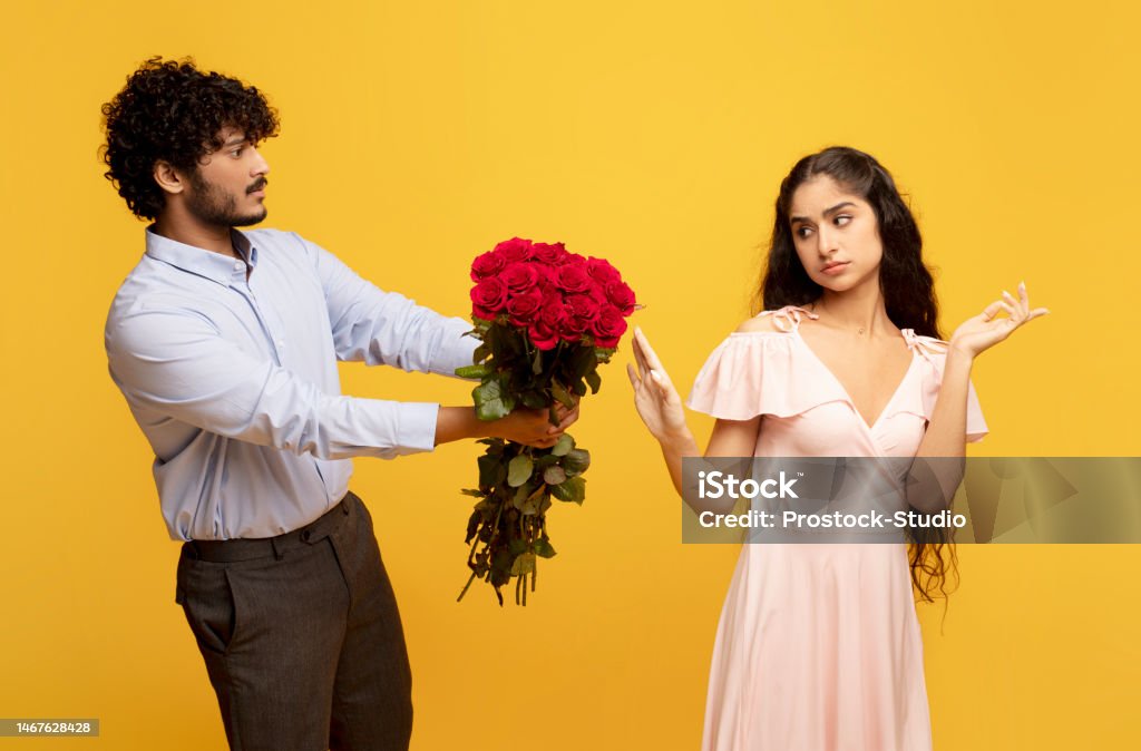 Unwanted confession. Displeased indian lady rejecting her admirer with roses on Valentine's Day, yellow background Unwanted confession. Displeased indian lady rejecting her admirer with roses on Valentine's Day, man giving flowers to his girlfriend, yellow studio background Rejection Stock Photo