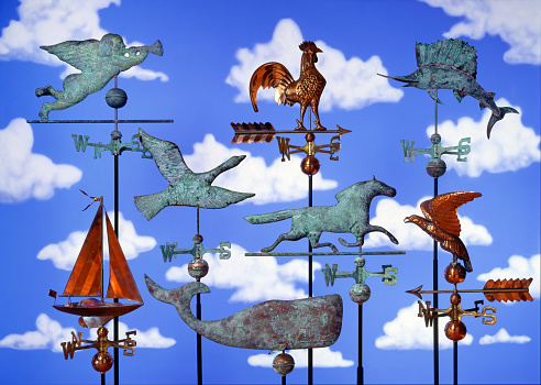 A collection of fanciful weather vanes in front of a hand painted cloud filled sky background.