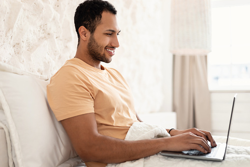 Side View Of Happy Arabic Male Freelancer Using Laptop Computer Working Online And Browsing Internet Sitting In Bedroom At Home. Freelance And Remote Job