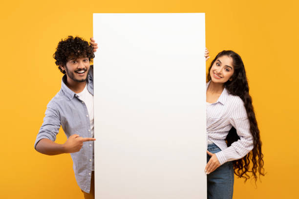 istock Happy indian couple in love standing by white empty board for advertisement or text over yellow studio background 1467632189
