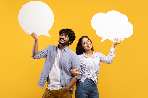 Portrait of young indian couple holding empty speech bubbles with free space for your text on yellow studio background. Millennial guy and his girlfriend posing with blank word clouds