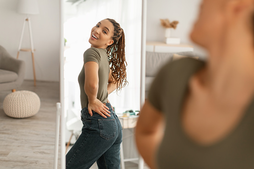 Great Weight Loss. Joyful African American Woman Looking At Her Reflection In Mirror After Successful Slimming And Diet Posing Standing At Home. Beauty And Healthcare Concept. Selective Focus