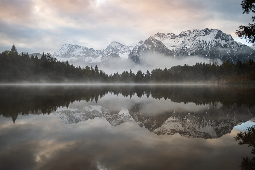 Lake Luttensee in Mittenwald, Bavaria is a breathtaking sight to behold. The still waters of the lake perfectly reflect the towering Karwendel Mountains in the background. The scene is made even more magical by the presence of a mysterious fog emerging from the water's surface, adding an otherworldly quality to the peaceful atmosphere. The result is a serene and beautiful moment frozen in time, a true masterpiece of nature's design.