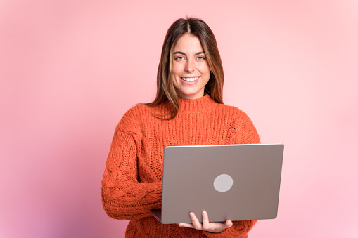 Positive young female in warm sweater smiling and looking at laptop screen while standing on pink background