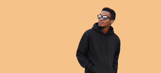 Portrait of stylish young african man looking away wearing black hoodie, sunglasses isolated on brown background, blank copy space for advertising text