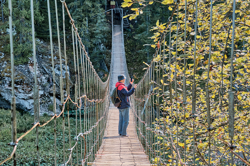 Senior male hiker with backpack taking pictures with cell phone on suspension bridge. Nature park Deer streams, Sverdlovsk region, Russia. Outdoor activities concept