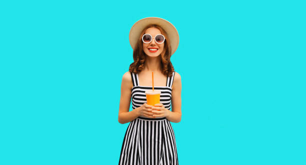 Summer portrait of beautiful young woman drinking fresh juice wearing straw round hat, striped dress on blue background stock photo