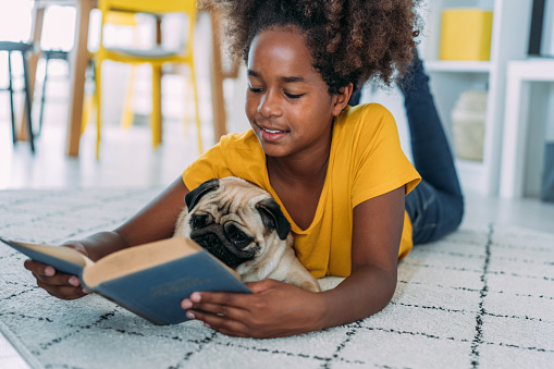 Shot of cute afro-american girl hugging her pug dog while reading a book on the floor in the living room at home.
