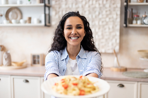 Happy latin woman showing spaghetti in the dish, stretching meal to camera and smiling, preparing delecious lunch at home, focus on positive housewife