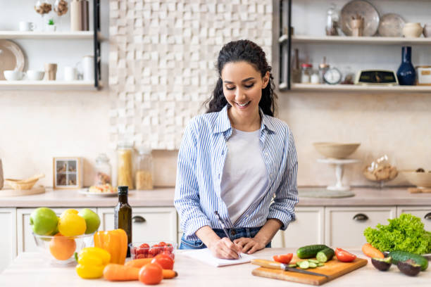 istock Happy young lady making notes on table with organic vegetables, cooking dinner in light kitchen interior 1467665171
