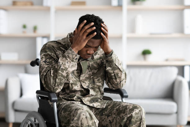 Upset black man sitting in wheelchair, wearing military uniform Upset young black man sitting in wheelchair, wearing military uniform, touching his head, suffering from loneliness and posttraumatic stress disorder, home interior, copy space depression land feature stock pictures, royalty-free photos & images