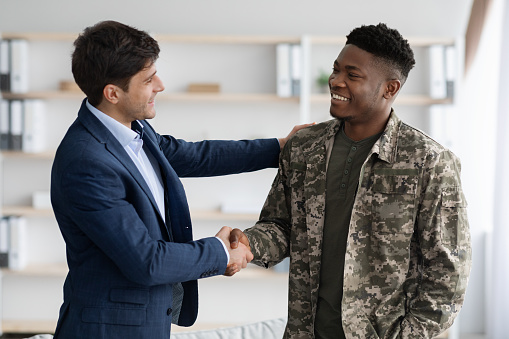 Cheerful guy in blue suit shaking happy young black military man hand, psychologist and soldier celebrating successful therapy, clinic interior. Military rehabilitation service concept