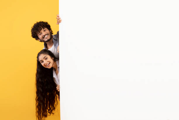 istock Excited indian couple standing behind white advertisement board and peeking out, showing empty space for your offer 1467665221