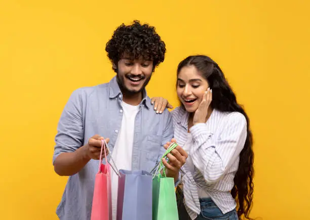 Photo of Shopaholics. Overjoyed indian couple holding shopping bags, looking inside, excited about their new purchases