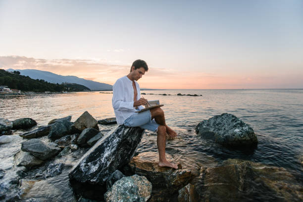 A male artist looking for inspiration by the sea. Sitting on the rock and painting by the sea at sunset under the mountain