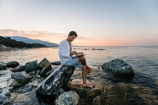A male artist looking for inspiration by the sea. Sitting on the rock and painting by the sea at sunset under the mountain