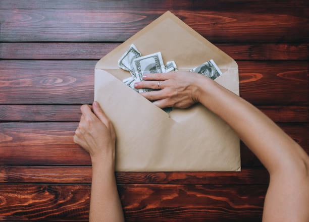 istock Two caucasian female hands hold an open beige craft envelope and take dollars on a brown wooden background. The concept of donation, alimony, winnings and cashback. Top view. 1467689044