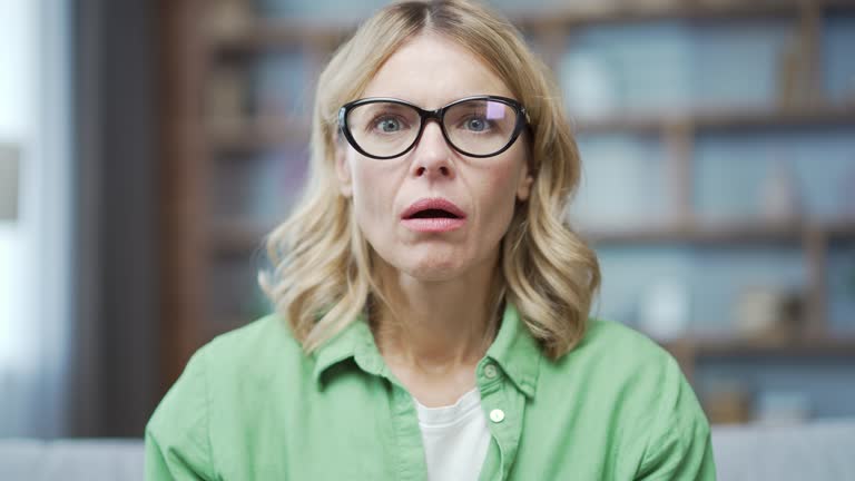 Webcam view. Close up of sad mature blonde woman looking surprised at camera or laptop screen while sitting in living room at home