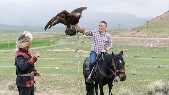 Issyk Kul, Kyrgyzstan - May 2022: Eagle trainer helping a tourist hold his golden eagle. Traditional tourism in Kyrgyzstan