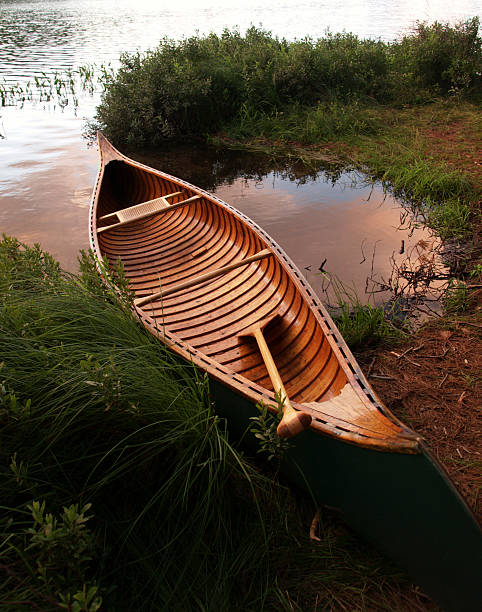 a wood and canvas canoe with paddle on lake shore in New York's Adirondack Park. Evening colors reflected in surface of the water.