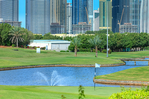 Golf fairways against the backdrop of contemporary architecture, in Dubai, United Arab Emirates in the Persian Gulf. In the background are tall buildings and modern architecture, in the area that is known as Jumeirah Lake Towers; the city is known for its skyscrapers and contemporary architecture. Some construction is still underway. There are also some villas in view in the background. Photo shot in the morning sunlight. Horizontal format; copy space. No people. Note to Inspector: The image was shot from a public road in the city.