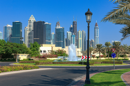 A typical example of urban landscaping in the modern Arabian city of Dubai, in the United Arab Emirates. In the background are skyscrapers of the area known as Jumeirah Lake Towers or JLT. In the foreground, date palms, flowering shrub and lawns. It is hard to believe that this is the Arabian desert; every blade of grass is artificially grown. In the background are also some villas by the roadside. Photo shot in the morning sunlight; horizontal format. Copy space. No people.