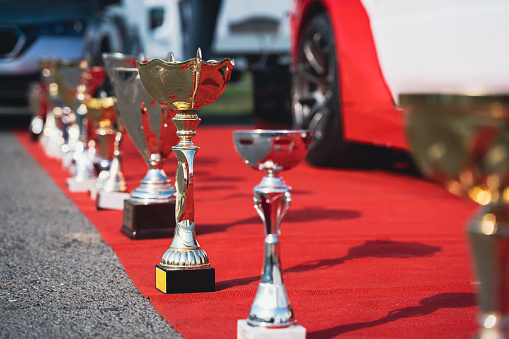 close up perspective view of golden and silver trophy standing  on display on a red carpet with racing cars in the background