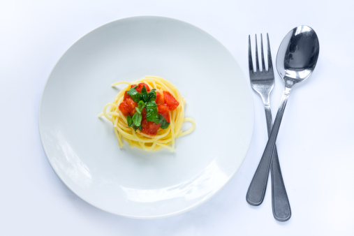 Small elegant or diet portion of pasta on a white plate on a light background. Fork and spoon are located next to the plate.