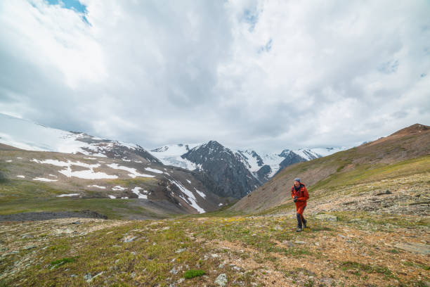 istock Man in red with camera walks with view to sunlit large snowy mountain range with sharp tops and glaciers under gray cloudy sky. Dramatic landscape with tourist in high mountains at changeable weather. 1467731813
