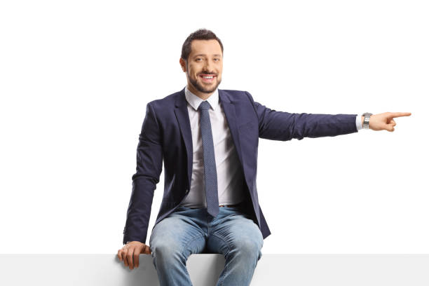istock Man in jeans and tie sitting on a blank panel and pointing to the side 1467751862