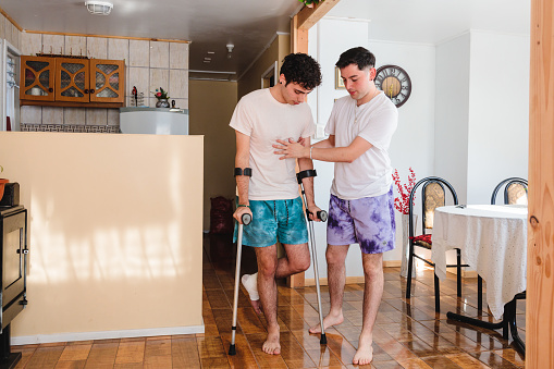 Man helps his gay partner who is using crutches inside their house. LGBT couples cohabitation routine