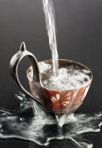 Old clay cup with water overflowing the edge, the water is blurred due to motion and the background is black/gray.