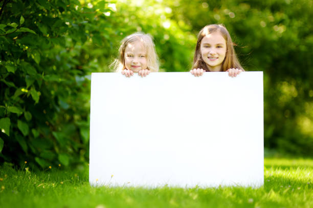 two cute little sisters holding big blank whiteboard on sunny summer day outdoors - preschooler childhood outdoors cheerful imagens e fotografias de stock