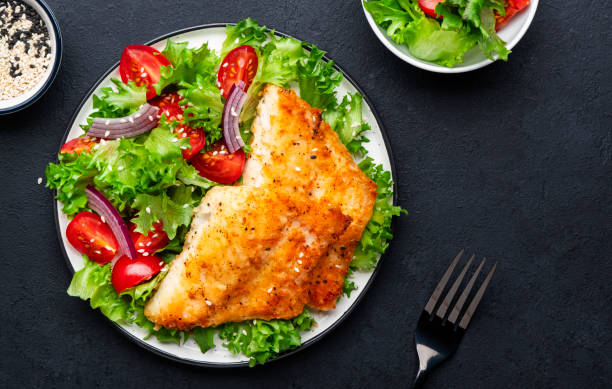istock Fried cod fish with salad garnish from lettuce, cherry tomatoes and red onion with sesame seeds, black table background, top view 1467788482