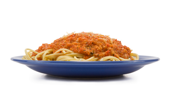 Top view of plate with spaghetti Bolognese pasta on wooden stand. There is fork and knife behind a plate. There is also bowl of parmesan cheese.