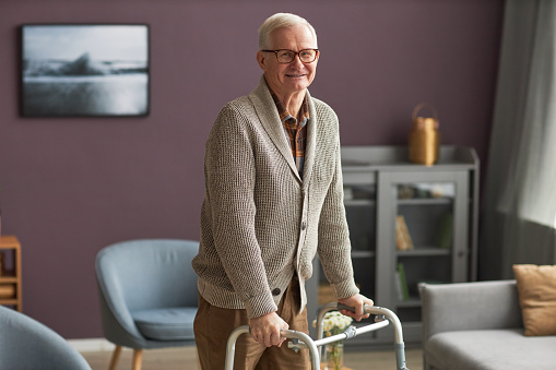 Portrait of elderly man with gray hair smiling at camera while walking with walker at home