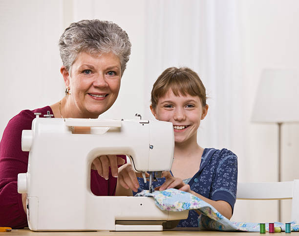 A young girl is using a sewing machine with her grandmother.  They are smiling at the camera.  Square framed shot.