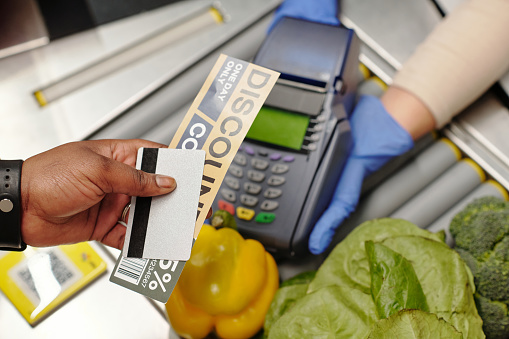 Overview of consumer hand holding credit card and discount coupon over payment terminal held by cashier in gloves during transaction