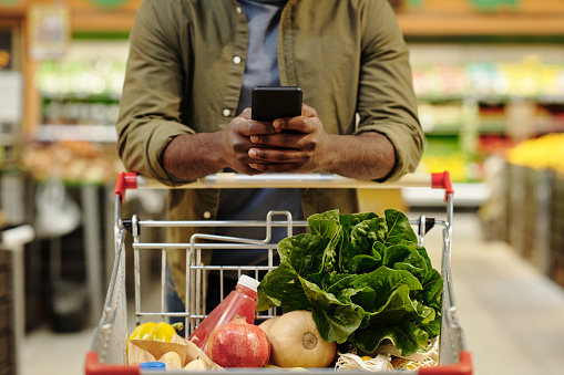 Young African American male consumer pushing shopping cart with fresh vegetarian food products and scrolling in smartphone in supermarket